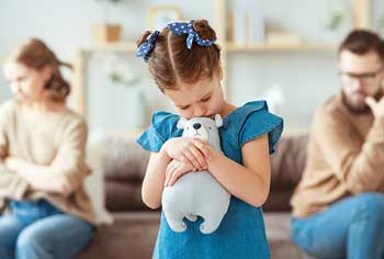 Child holding a teddybear with fighting parents sitting at oppposite ends of sofa in background